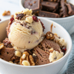 Scoops of rocky road vegan ice cream in a white bowl topped with marshmallows, chocolate chunks, and nuts.