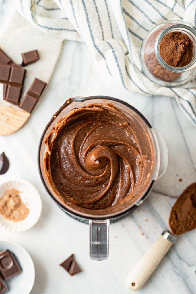 Blender with smooth banana-almond mixture surrounded by ingredients on a marble surface.