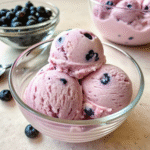 Three scoops of blueberry vegan ice cream in a glass bowl with fresh blueberries around.