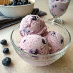 Scoops of creamy blueberry vegan ice cream in a glass bowl with fresh blueberries.