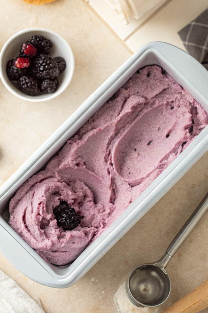 Blackberry ice cream mixture churned in a white loaf pan with a scoop and blackberries nearby