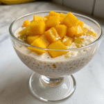 Glass cup filled with creamy mango sago topped with fresh mango cubes on a white table