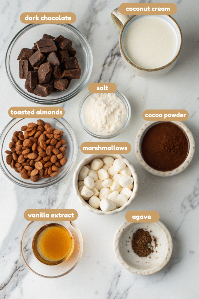 vegan rocky road ingredients in small bowls, including cocoa powder, maple syrup, chocolate chunks, almond milk, almonds, marshmallows, vanilla extract, and salt.