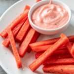 Fresh watermelon fries served with creamy pink dipping sauce on a white plate.