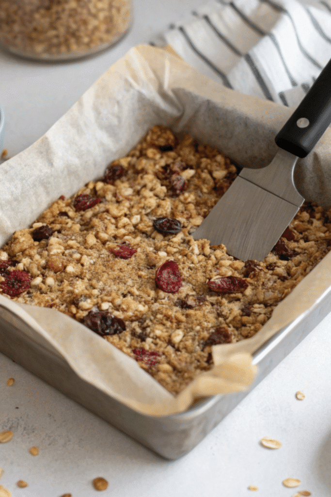 Pressing fruit granola bar mixture into a square pan lined with parchment paper, ready to set.