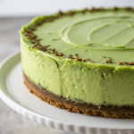 Whole green cheesecake with smooth matcha filling and nutty crust on a white cake stand