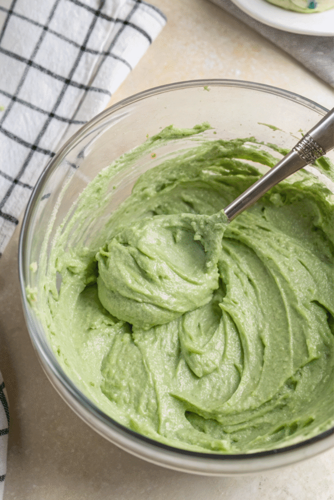Mixing smooth, vibrant green batter for Green Velvet Cheesecake in a glass bowl with a spoon, next to a white cloth with a dark gray checkered pattern.