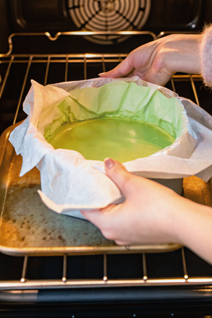 Hands placing a green matcha basque cheesecake batter into the oven inside a parchment-lined cake pan.