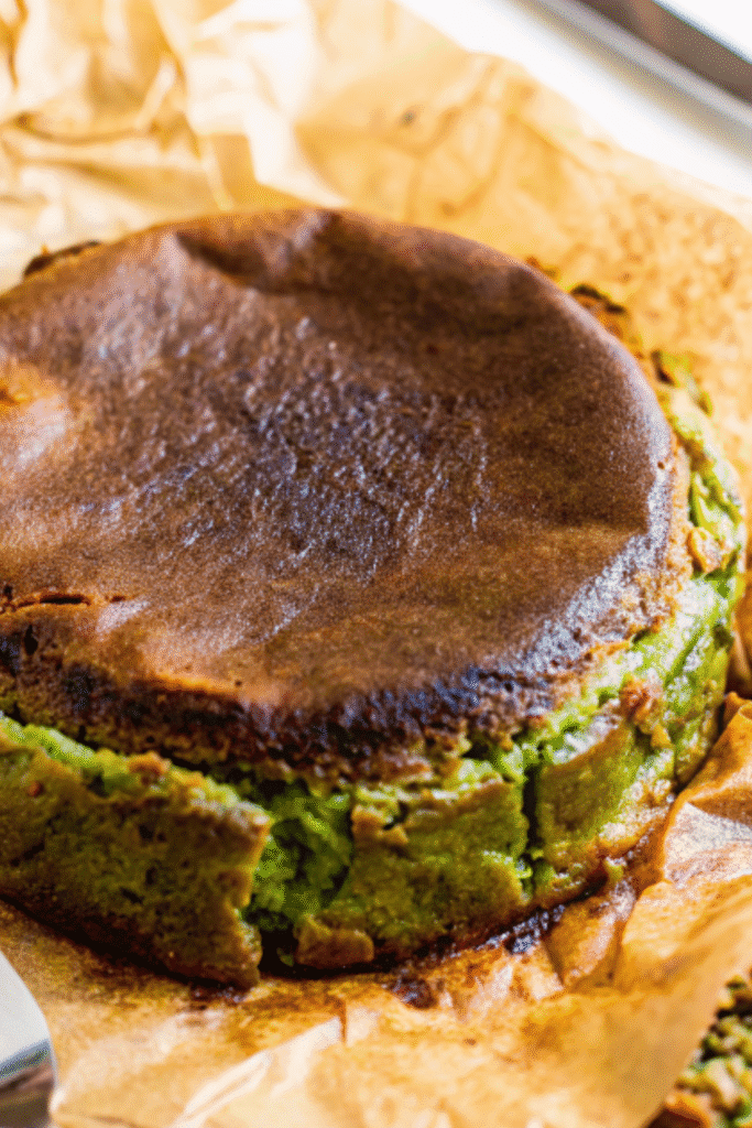 Side angle of a baked matcha cheesecake with parchment partially removed, revealing its vibrant green interior and browned top.