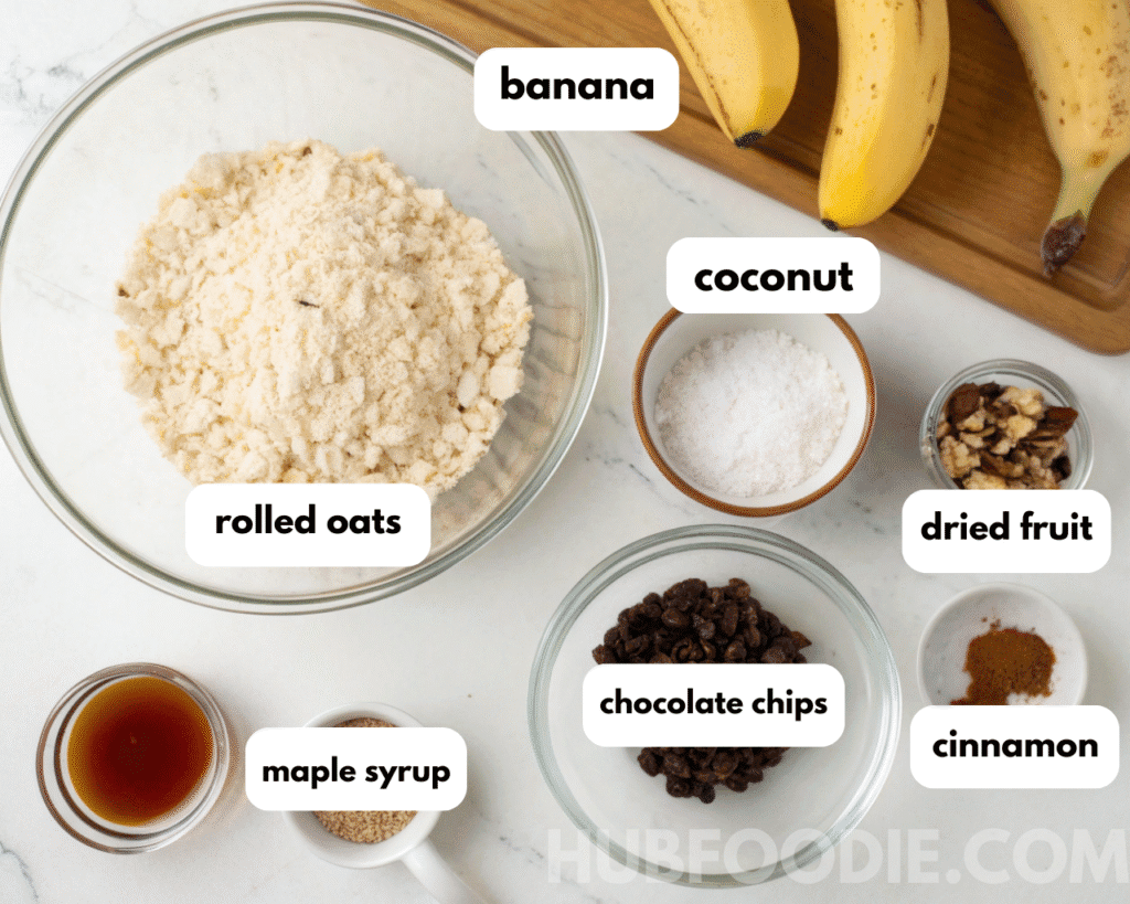 Nut free granola bars ingredients including rolled oats, banana, coconut, dried fruit, maple syrup, chocolate chips, cinnamon, and seeds arranged in glass bowls on a white kitchen counter.