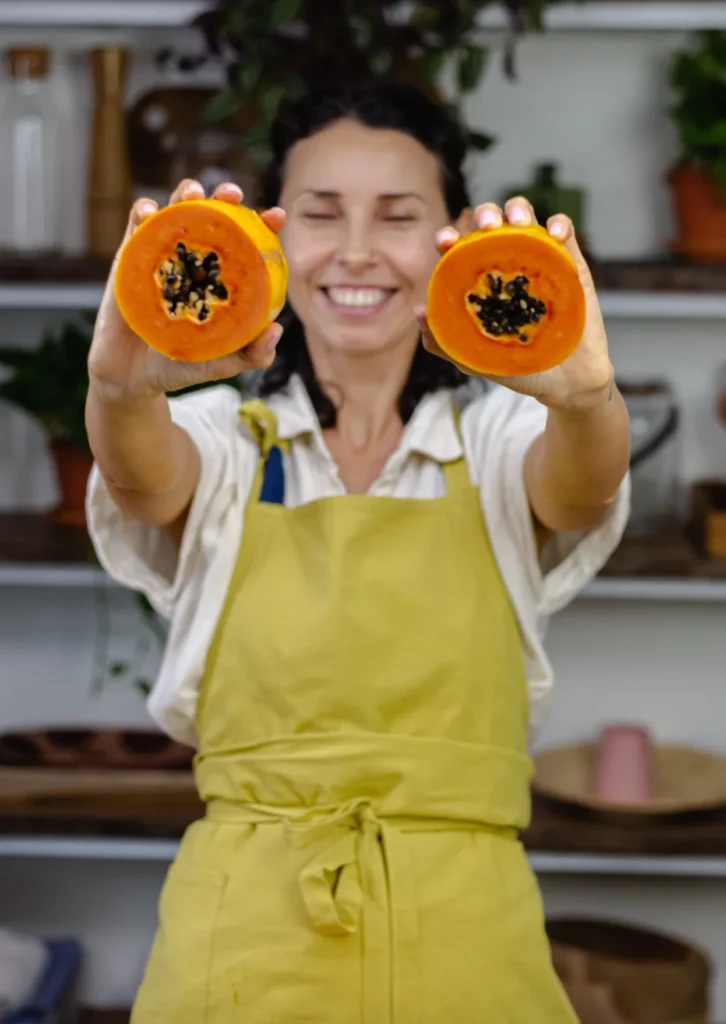 Smiling woman in yellow apron holding two halves of a papaya towards the camera.