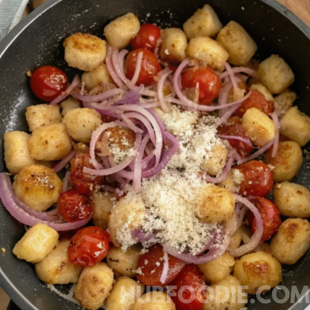 Crispy gnocchi in a skillet topped with red onions, cherry tomatoes, grated cheese, and herbs.